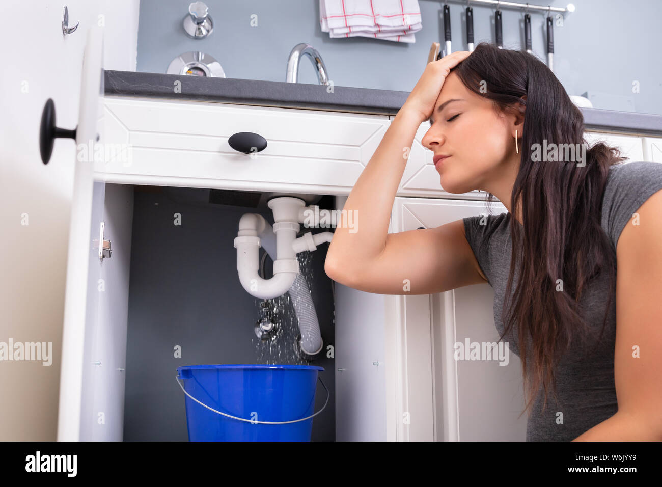 Worried Woman Collecting Water Leaking From Sink In Blue Bucket Stock Photo
