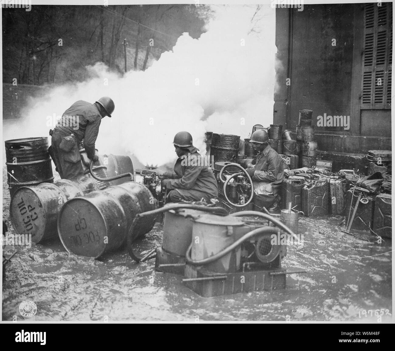 Soldiers of the 161st Chemical Smoke Generating Company, U.S. Third Army, move a barrel of oil in preparation to refilling an M-2 smoke generator, which spews forth a heavy cloud of white smoke. These men are engaged in laying a smoke screen to cover bridge building activities across the Saar River near Wallerfangen, Germany., 12/11/1944 Stock Photo
