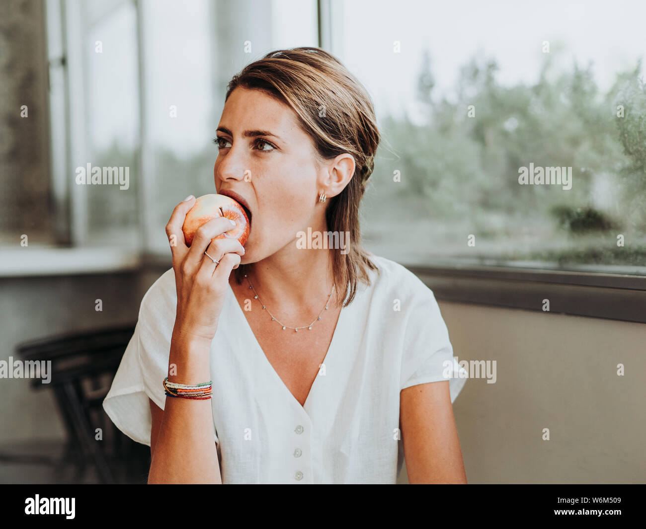 Young woman eating an apple by biting in front of the big window of the kitchen of her house. Stock Photo