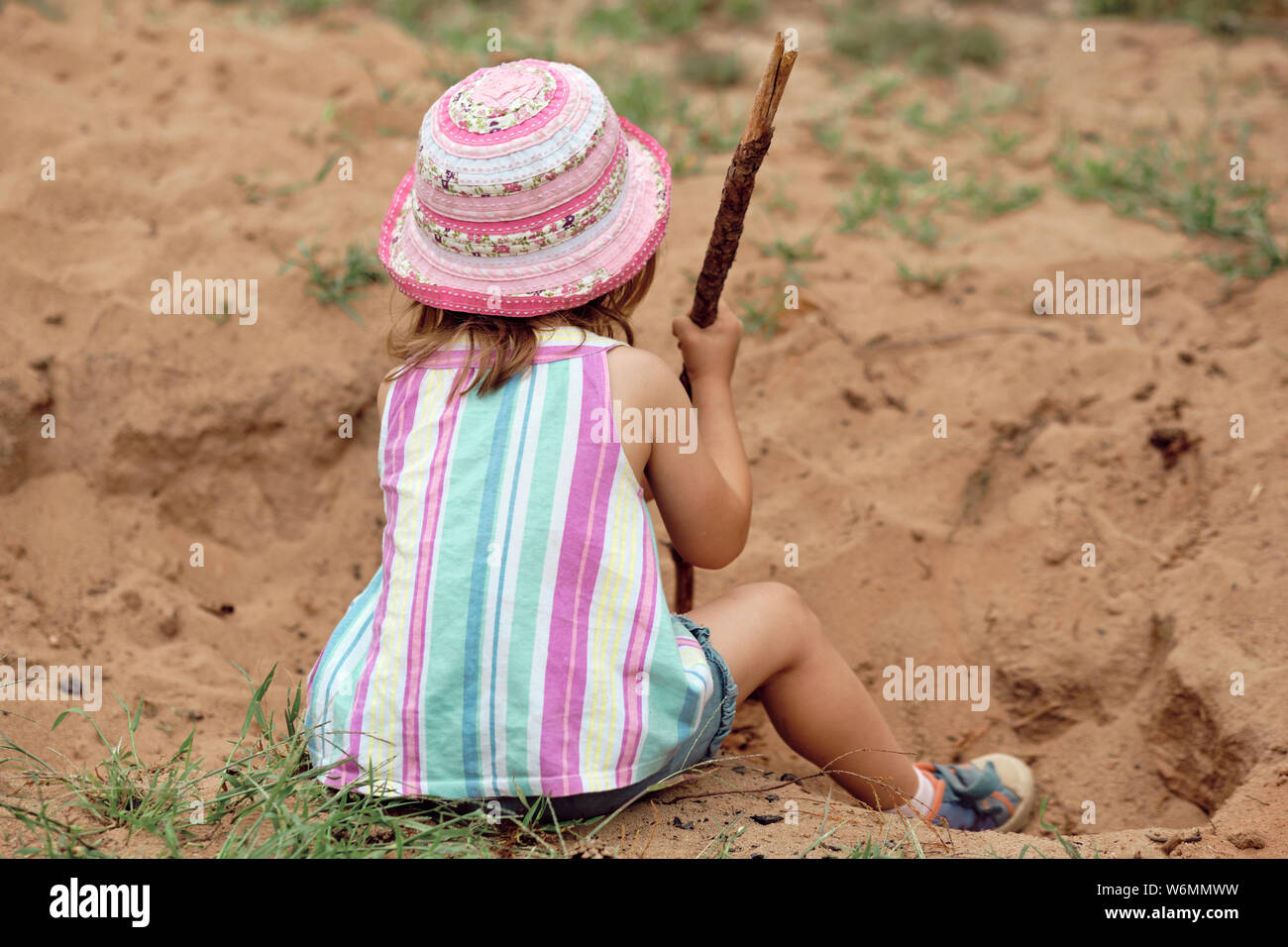 A child girl of 3-4 years old in summer clothing sitting and digging in the sand with a wooden stick Stock Photo