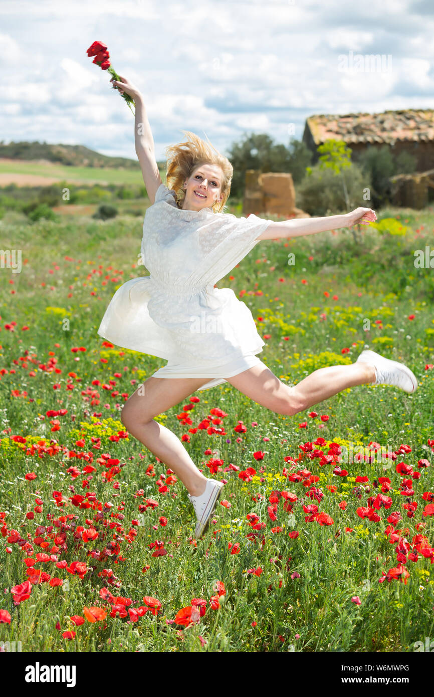 Jolly young female in white dress jumping in poppy field of wild