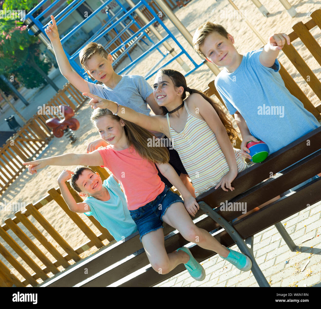 Five glad children sitting on a bench at the playground and point to ...