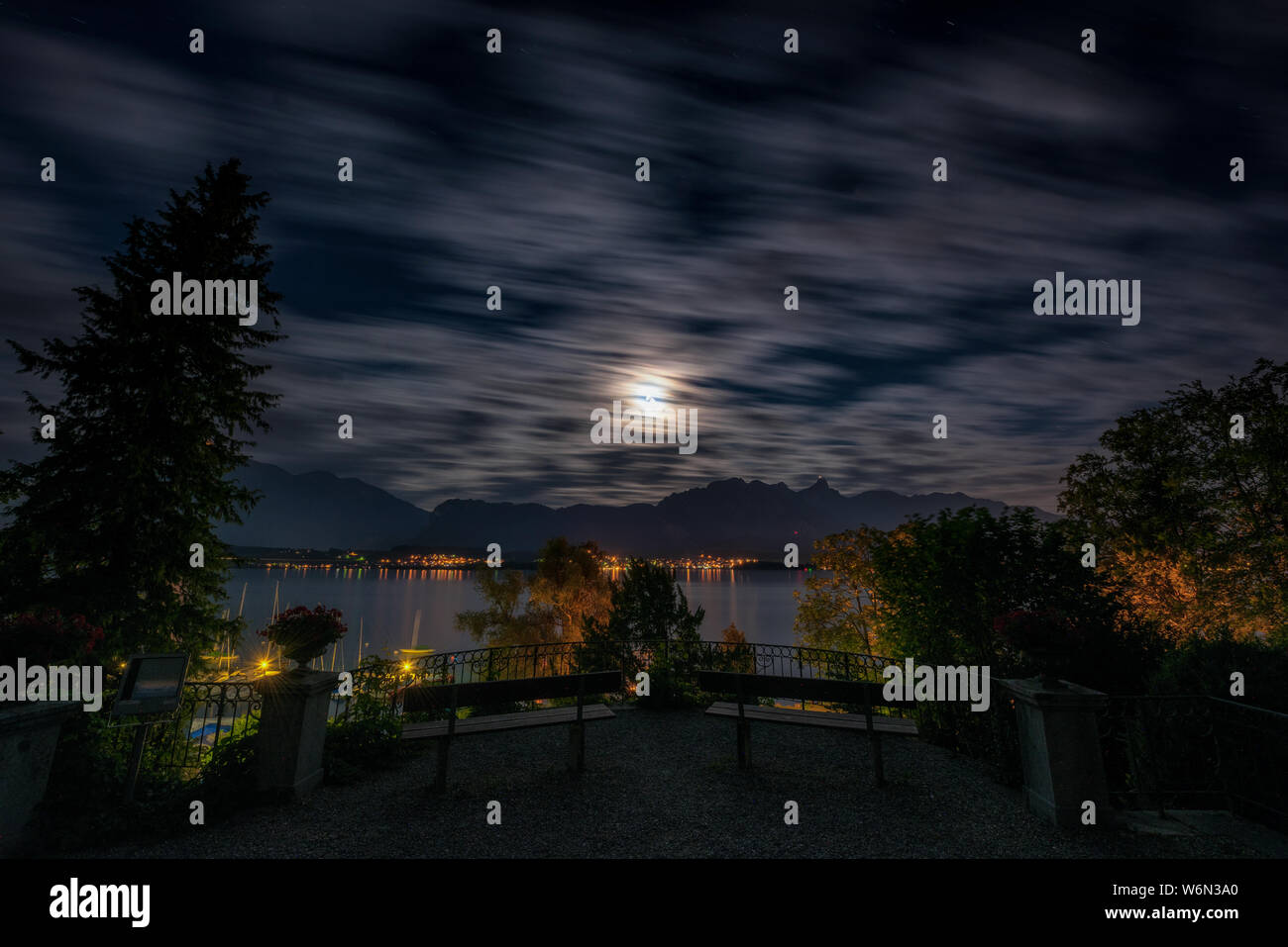 long exposure of a fringed night sky with moonlight over the Stockhorn mountain range, Lake Thun, Hilterfingen marina and two benches and railing of the Hünegg castle park Stock Photo