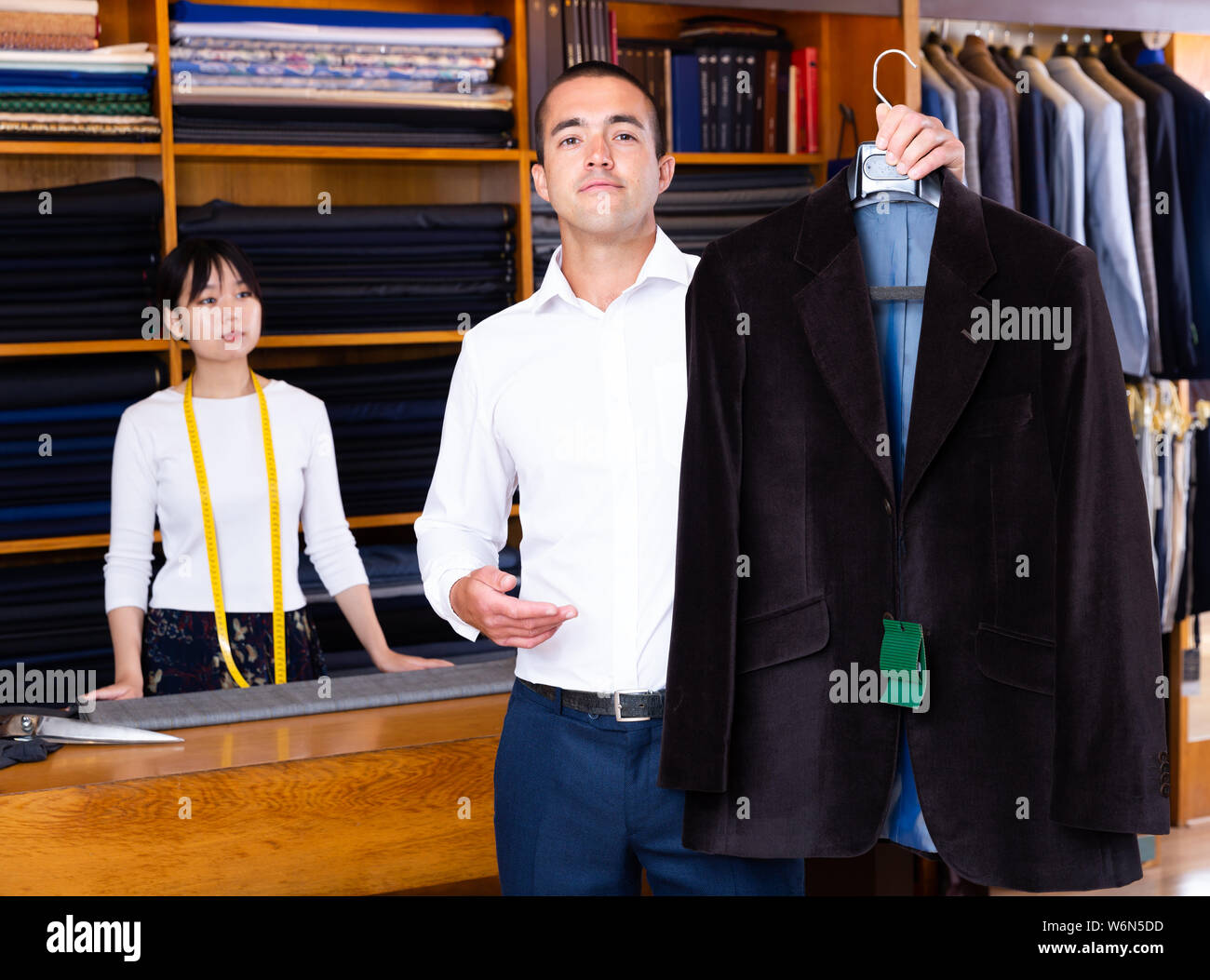 Satisfied male client of sewing workshop showing his new suit jacket on background of young Chinese female tailor Stock Photo