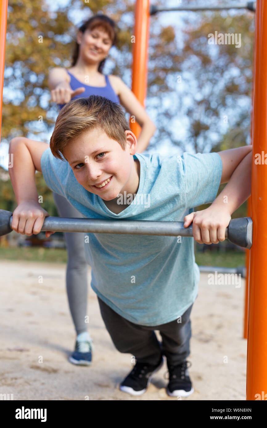 Preteen boy doing push-ups on gymnastics bar during workout with his mother on outdoor sports ground Stock Photo