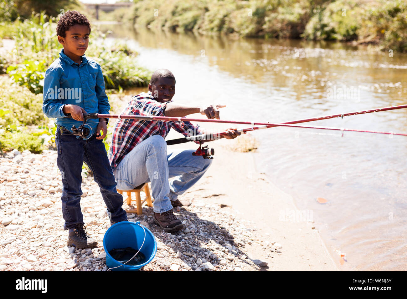 Portrait of cheerful afro man and his little son fishing with rods on river Stock Photo
