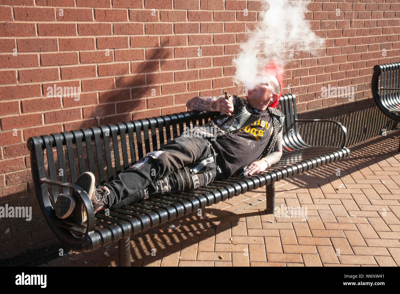 Punk rocker with red spiked mohican haircut enjoying a smoke in Blackpool, Lancashire, UK. Aug, 2019. Lee Talbot from Leeds at the Rebellion Festival world's largest punk festival in Blackpool. At the beginning of August, Blackpool's Winter Gardens plays host to a massive line up of punk bands for the 21st edition of Rebellion Festival attracting thousands of tourists to the resort. Stock Photo