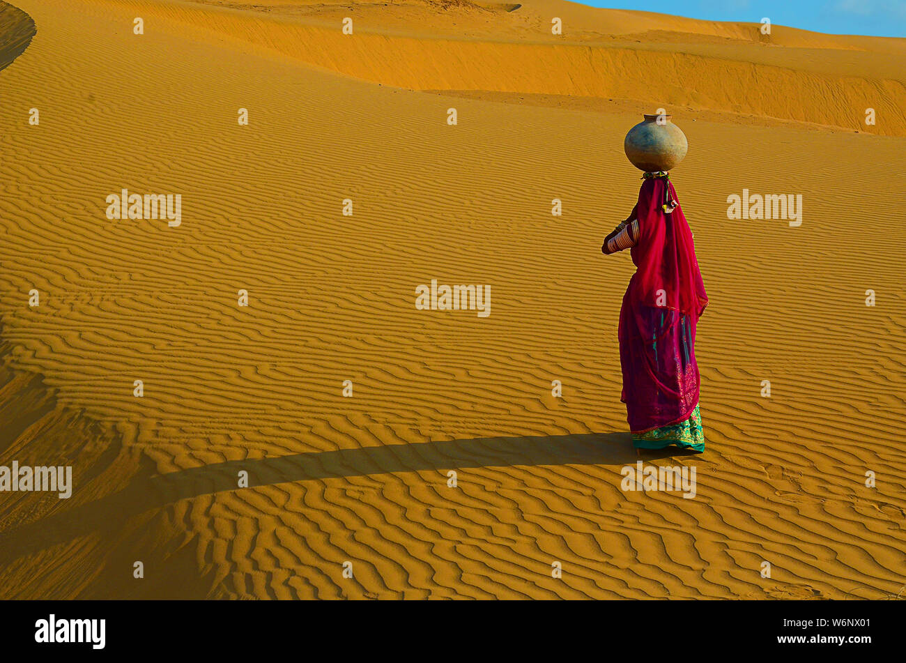 Indian woman carrying heavy jug of water on her head and walking on a yellow sand dune in the hot summer desert against blue sky.water crises, jaisalm Stock Photo