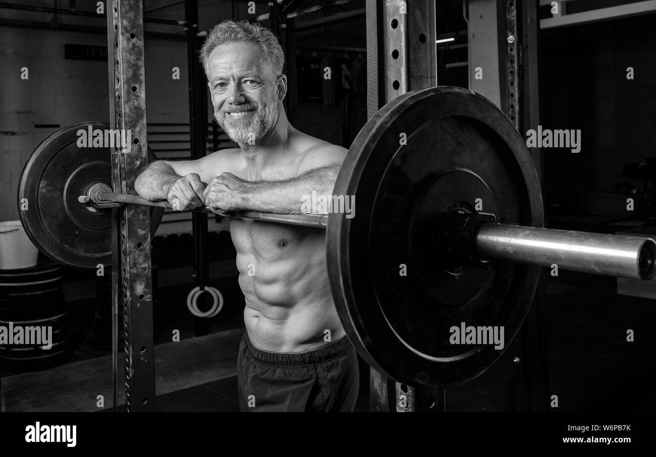 Black and white portrait of an attractive middle aged athlete with Sixpack. The athletic and smiling bearded man is leaning against the barbell rack. Stock Photo