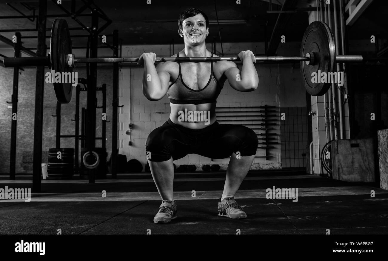 A fit short haired woman with Sixpack is doing weight lifting with a barbell in a gym. The muscular woman is doing front squats. Stock Photo