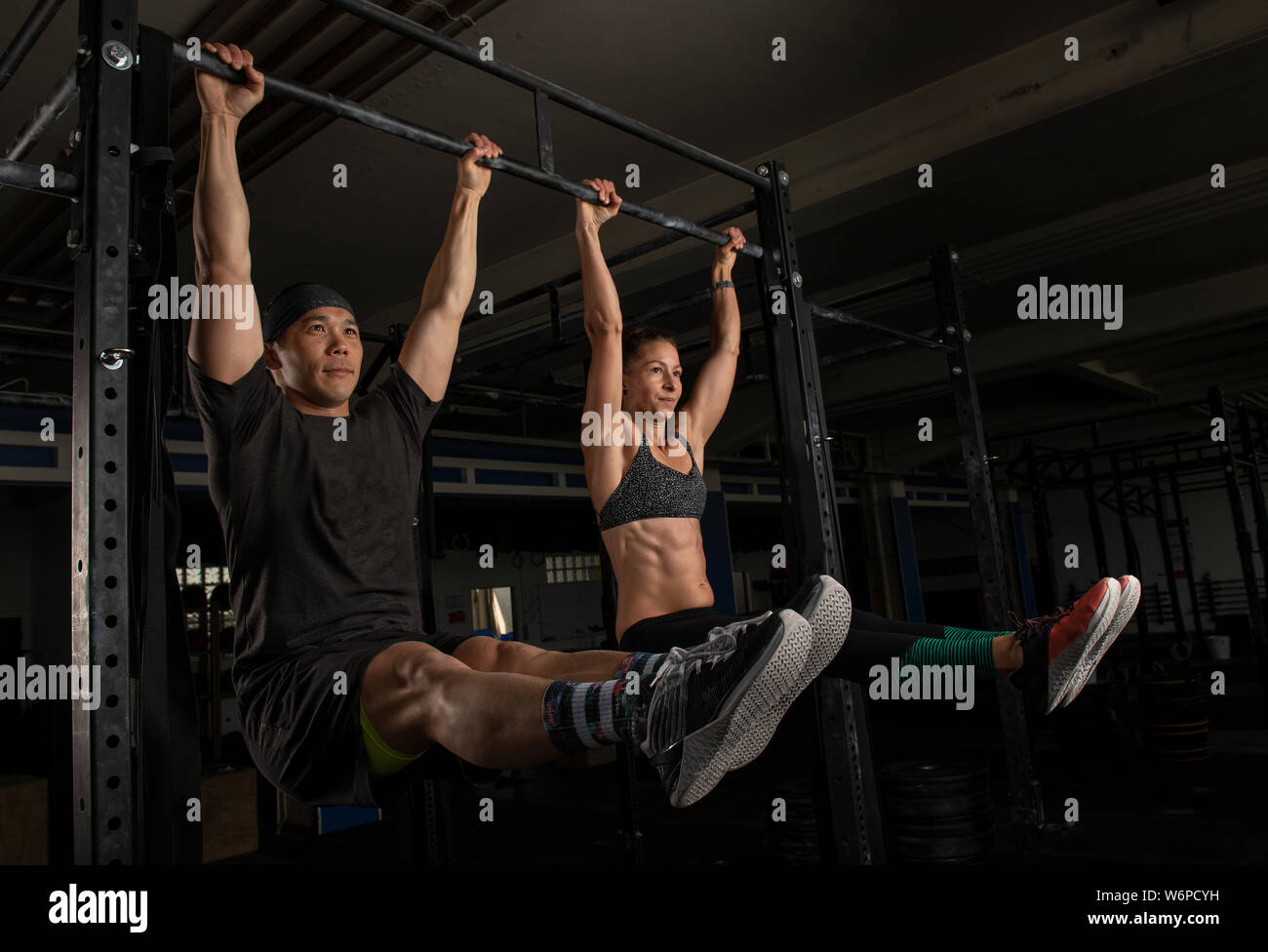 A muscular young couple is doing a fitness training together and having fun. Strong man and woman are doing the exercise l sit on the horizontal bar. Stock Photo