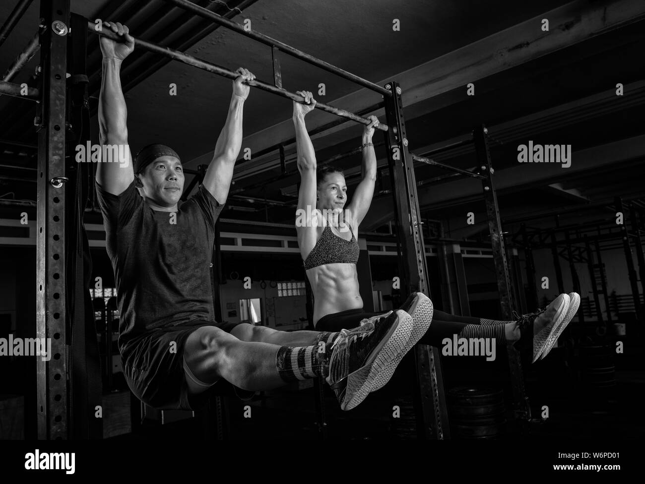 An athletic young couple is doing a fitness training together and having fun. Strong man and woman are doing the exercise l sit on the horizontal bar. Stock Photo