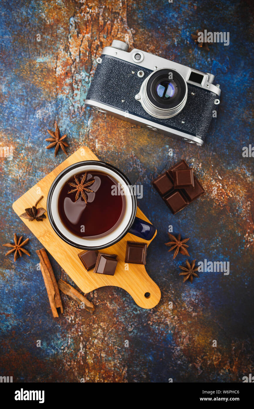 Blue enamelled cup of tea, cinnamon sticks, anise stars, pieces of dark chocolate on a wooden cutting board and a retro camera on a dark background. T Stock Photo