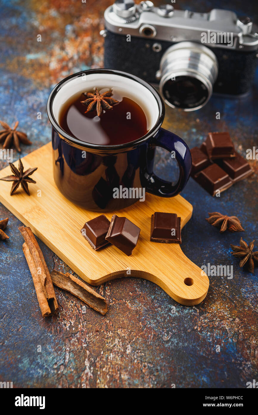 Blue enamelled cup of tea, cinnamon sticks, anise stars, pieces of dark chocolate on a wooden cutting board and a retro camera on a dark background Stock Photo