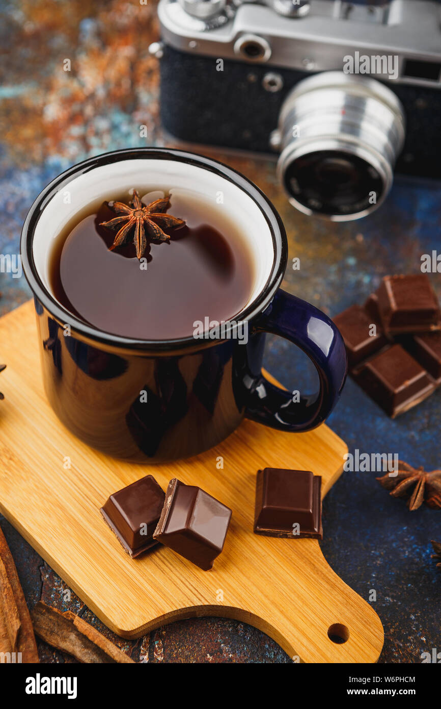 Blue enamelled cup of tea, cinnamon sticks, anise stars, pieces of dark chocolate on a wooden cutting board and a retro camera on a dark background Stock Photo