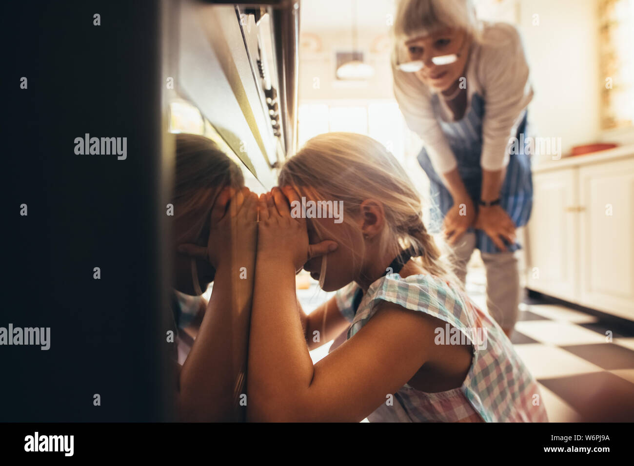 Close up of a girl looking inside an oven through the glass door. Granny and kid in kitchen looking for cake inside the oven. Stock Photo