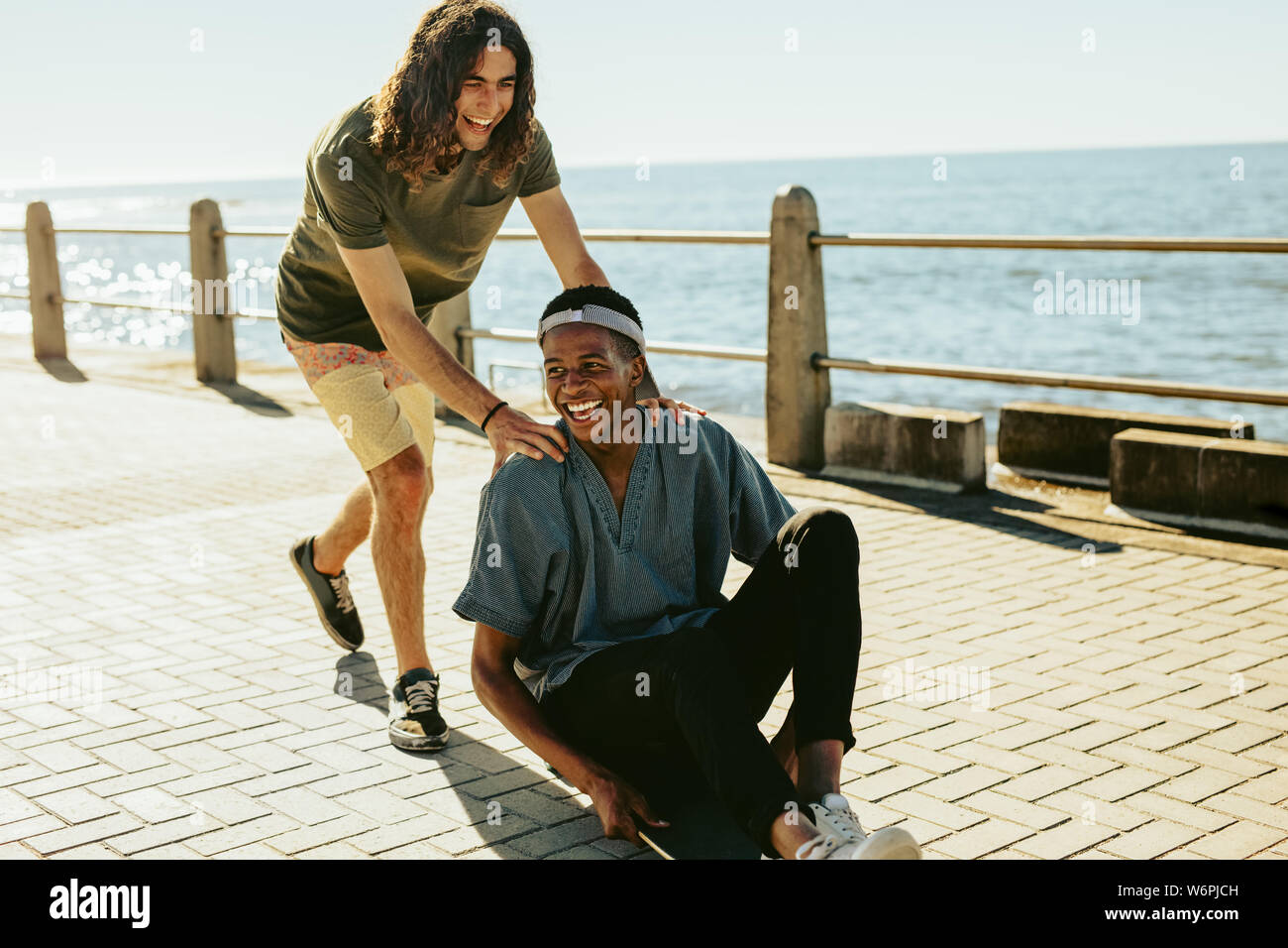 Young man being pushing on skateboard by his friend outdoors on road by the sea. Male friends having fun outdoors on a summer day. Stock Photo