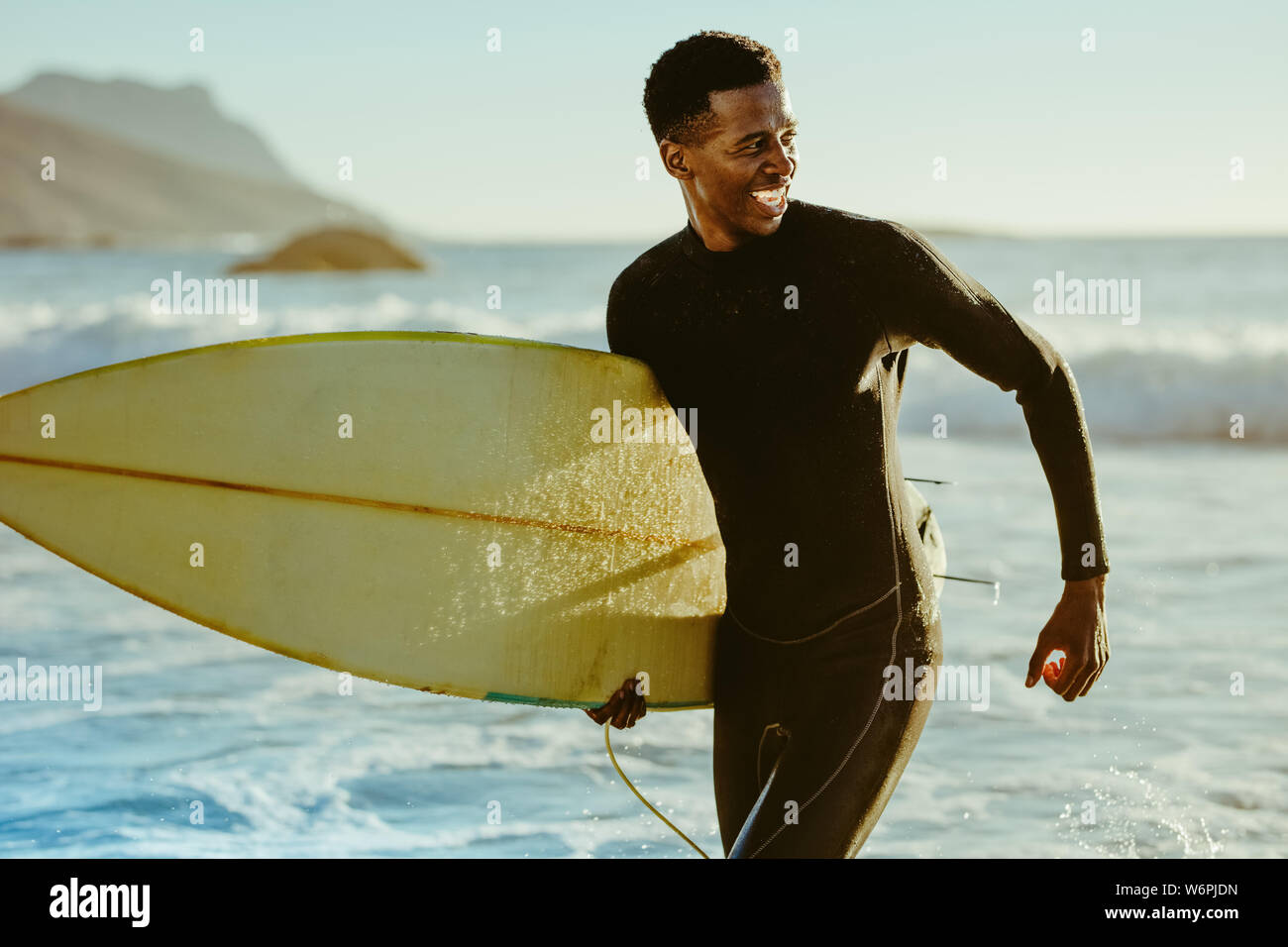 Smiling african male running out of the ocean after water surfing. Happy young man with surfboard on the beach enjoying holidays. Stock Photo