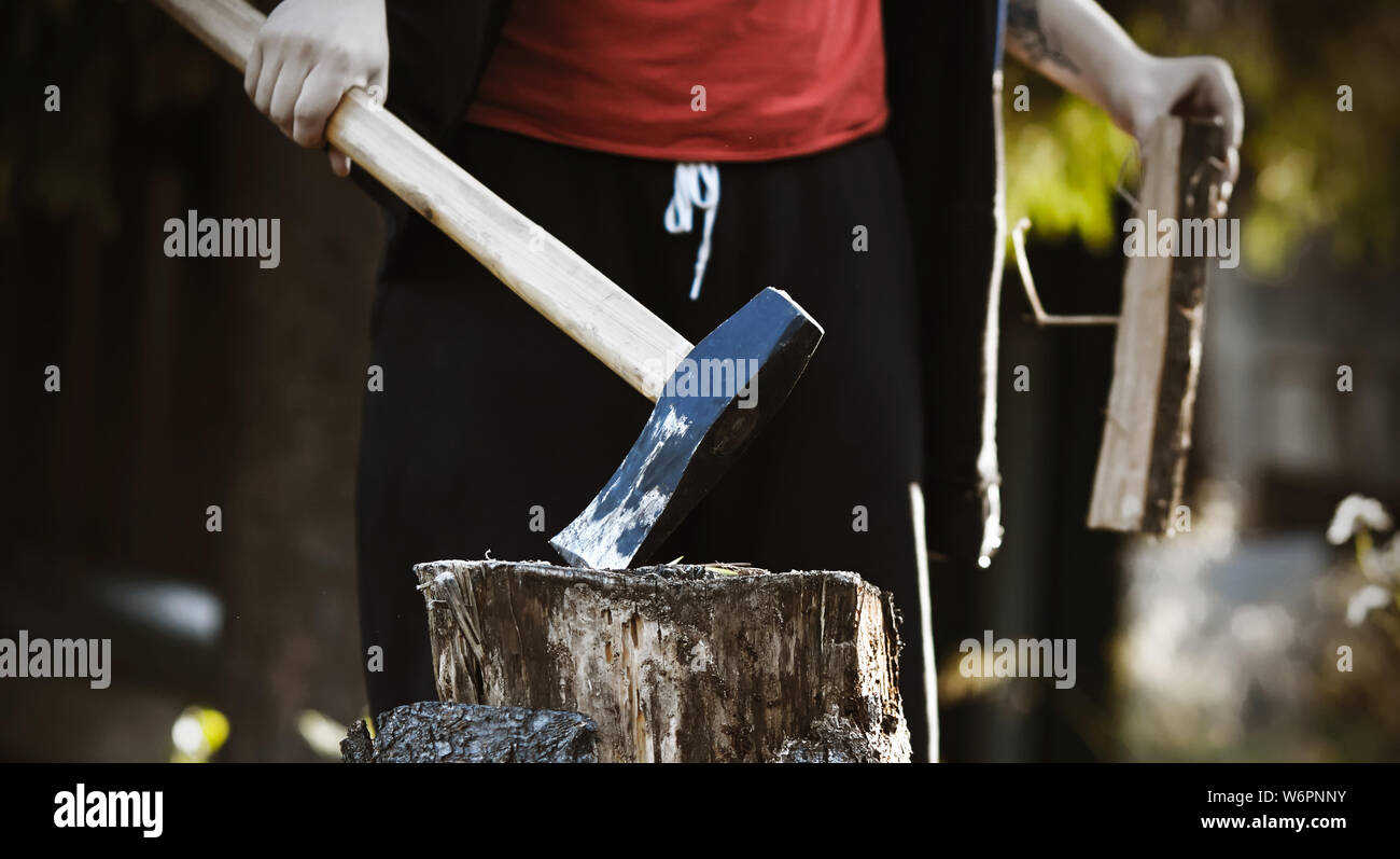 The woodcutter guy , dressed in black clothes and a red t-shirt, holds a sharp long axe in one hand, sticking it into a stump, and in the other hand h Stock Photo