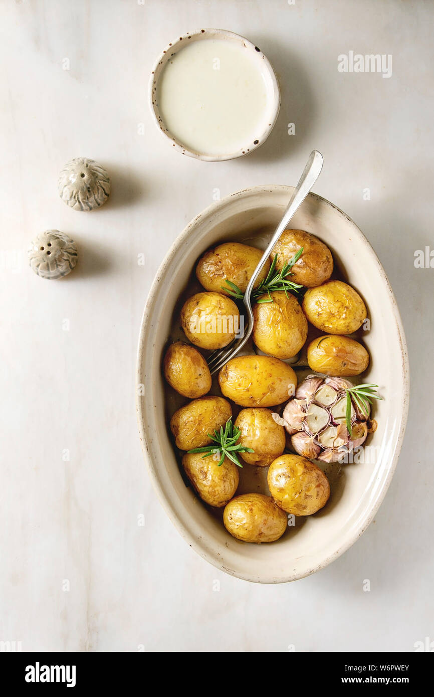 Young baked potatoes with garlic and rosemary in ceramic baking dish, served with cheese sauce, salt and pepper over white marble background. Flat lay Stock Photo