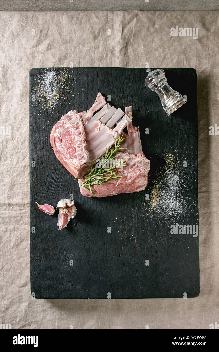 Raw uncooked rack of lamb on black burned wooden cutting board with salt, herbs rosemary, pepper and garlic over grey table cloth as background. Flat Stock Photo