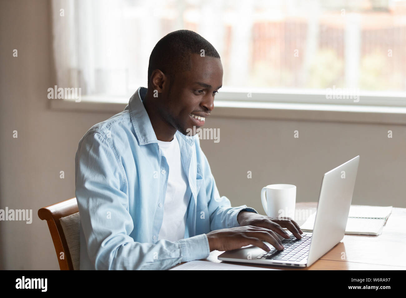 African millennial guy sitting at table typing on laptop Stock Photo
