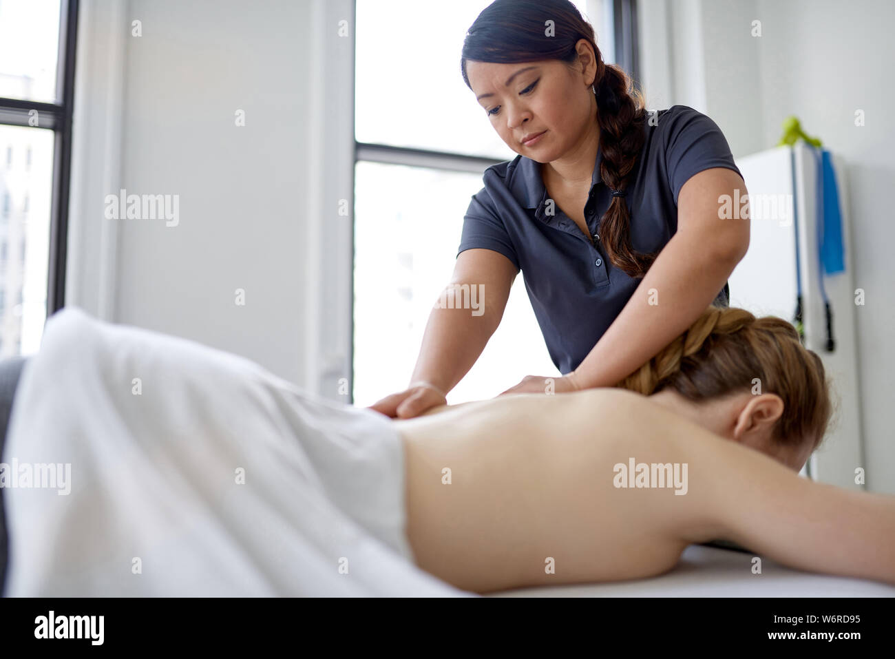 Chinese woman massage therapist giving a treatment to an attractive blond client on massage table in a bright medical office Stock Photo