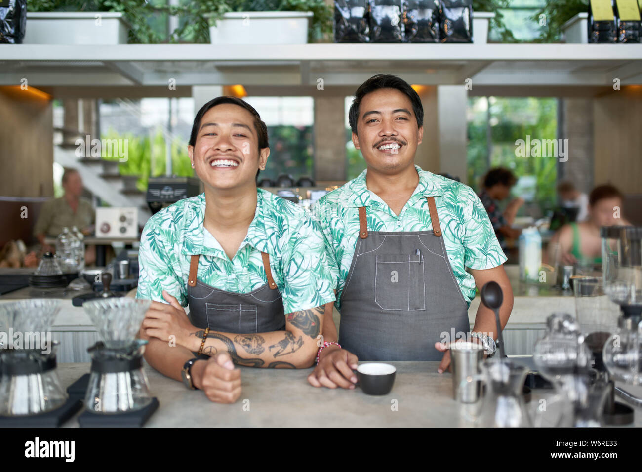 Lifestyle portrait of two friendly smiling balinese millennial baristas wearing trendy clothing and aprons in hipster cafe serving fair-trade coffee Stock Photo