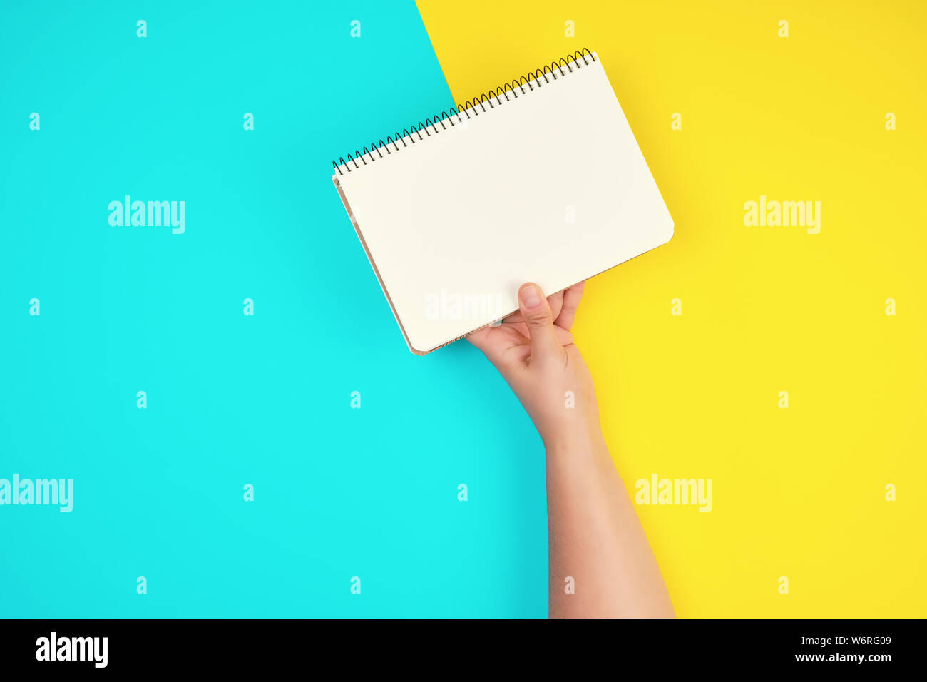 woman's hand holds an open spiral notebook with blank white sheets on a yellow-blue background Stock Photo