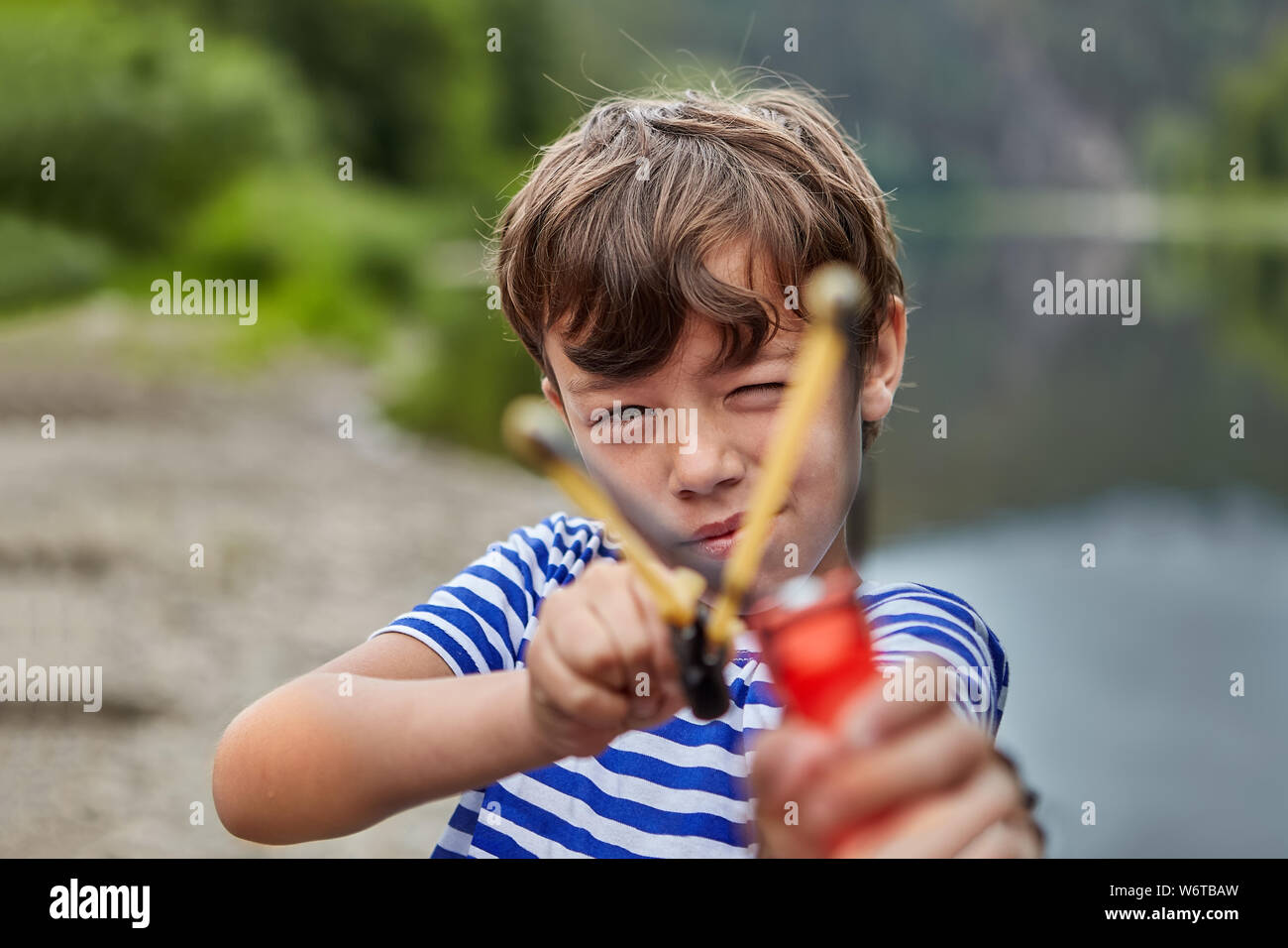 Little white boy is standing in forest or pakr with slingshot in his hands and preparing for shooting strewing up his eyes, boy is aiming. Stock Photo
