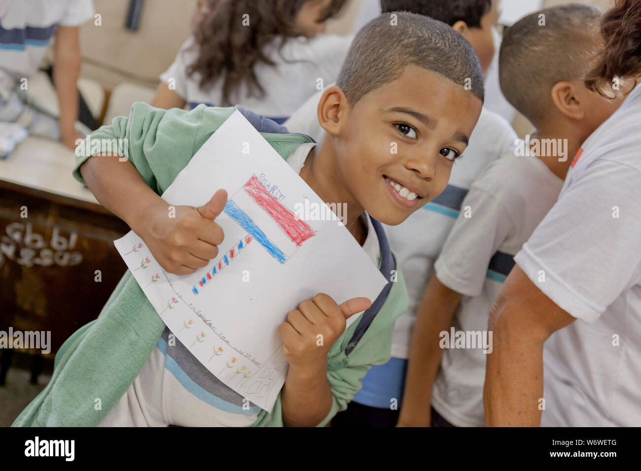 Young Brazilian school boy holding a drawing he made smiling and putting two thumbs up at the camera with his peers in the background Stock Photo