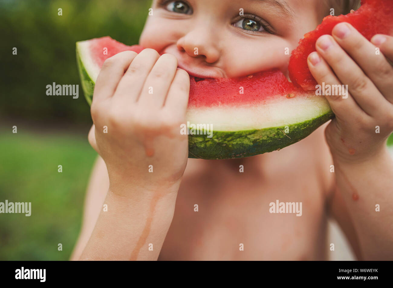 A young child is taking a bite out of a big slice of watermelon and looking directly into the camera. Stock Photo