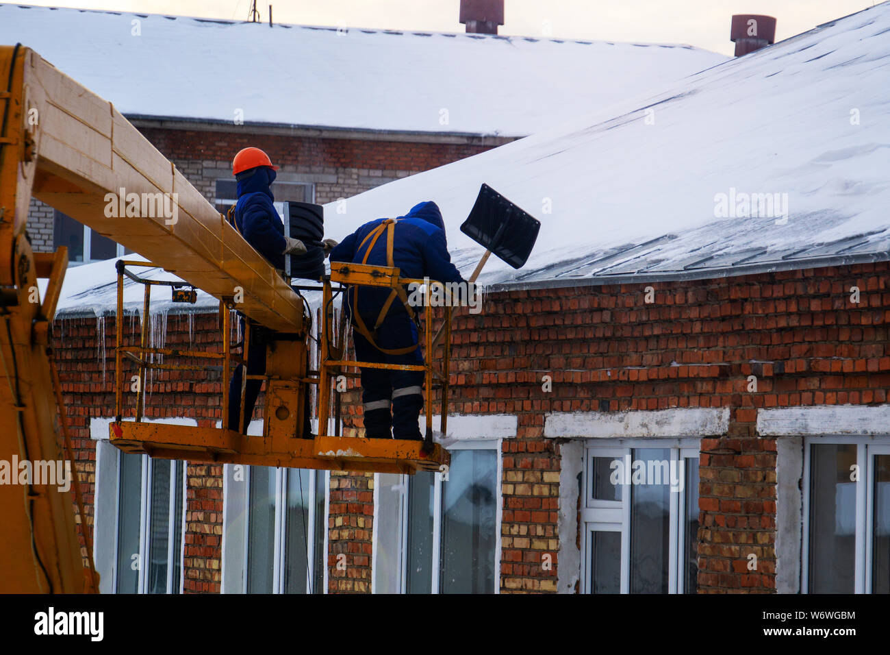Workers in overalls and orange helmets on the crane basket remove icicles from roof of the house on a winter day - cleaning the roofing, utility servi Stock Photo
