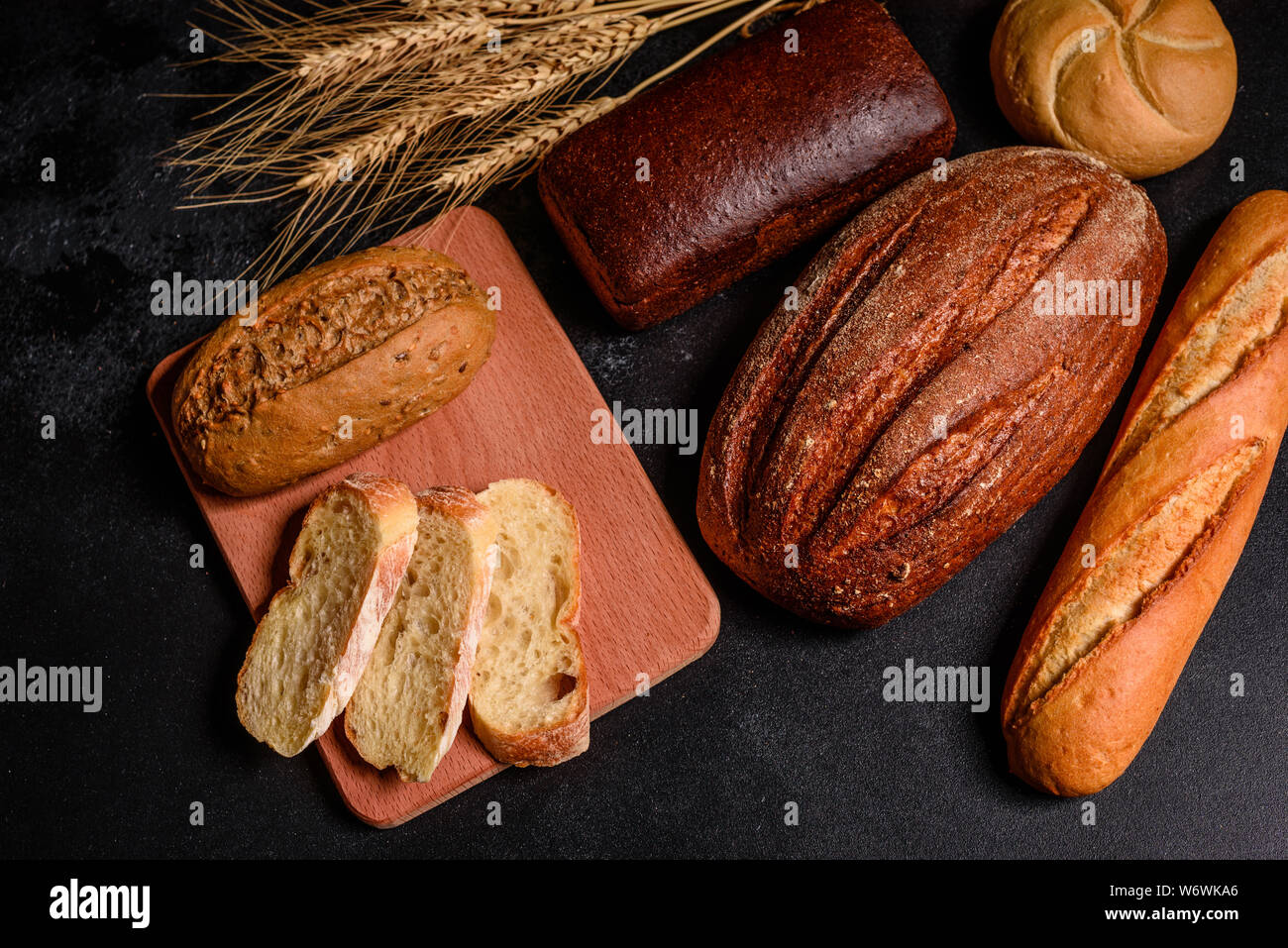 Fresh fragrant bread with grains and cones of wheat against a dark background. Assortment of baked bread on wooden table background Stock Photo