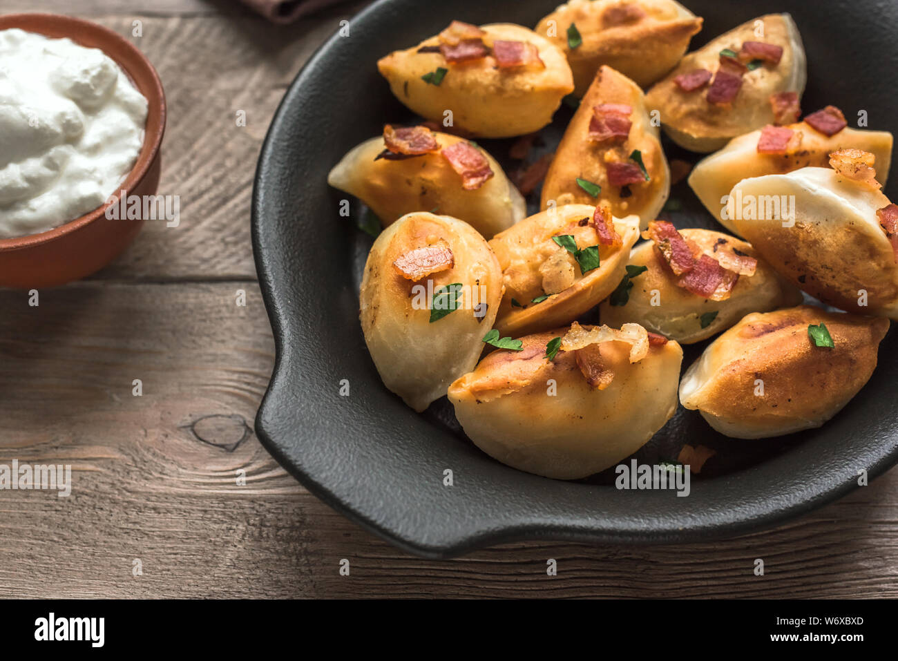 Fried dumplings stuffed with potato and meat (pierogi) sprinkled with bacon and  parsley and sour cream on wooden table, close up. Stock Photo