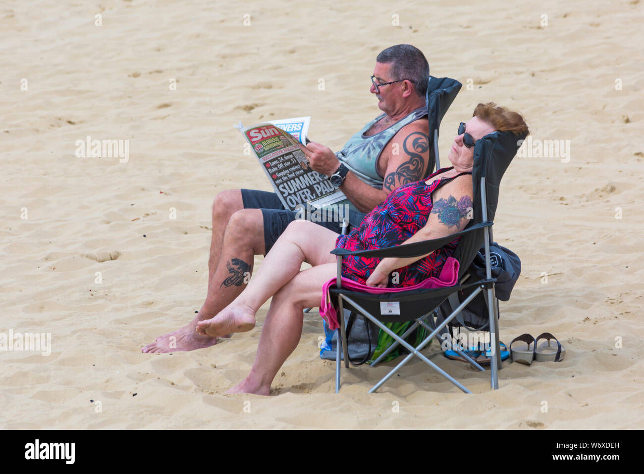 Bournemouth, Dorset UK. 3rd Aug 2019. UK weather: overcast and cloudy, but warm and muggy. Beach goers head to the beaches at Bournemouth to enjoy the warm weather. Couple relaxing in chairs on the beach, man reading the Sun newspaper. Credit: Carolyn Jenkins/Alamy Live News Stock Photo