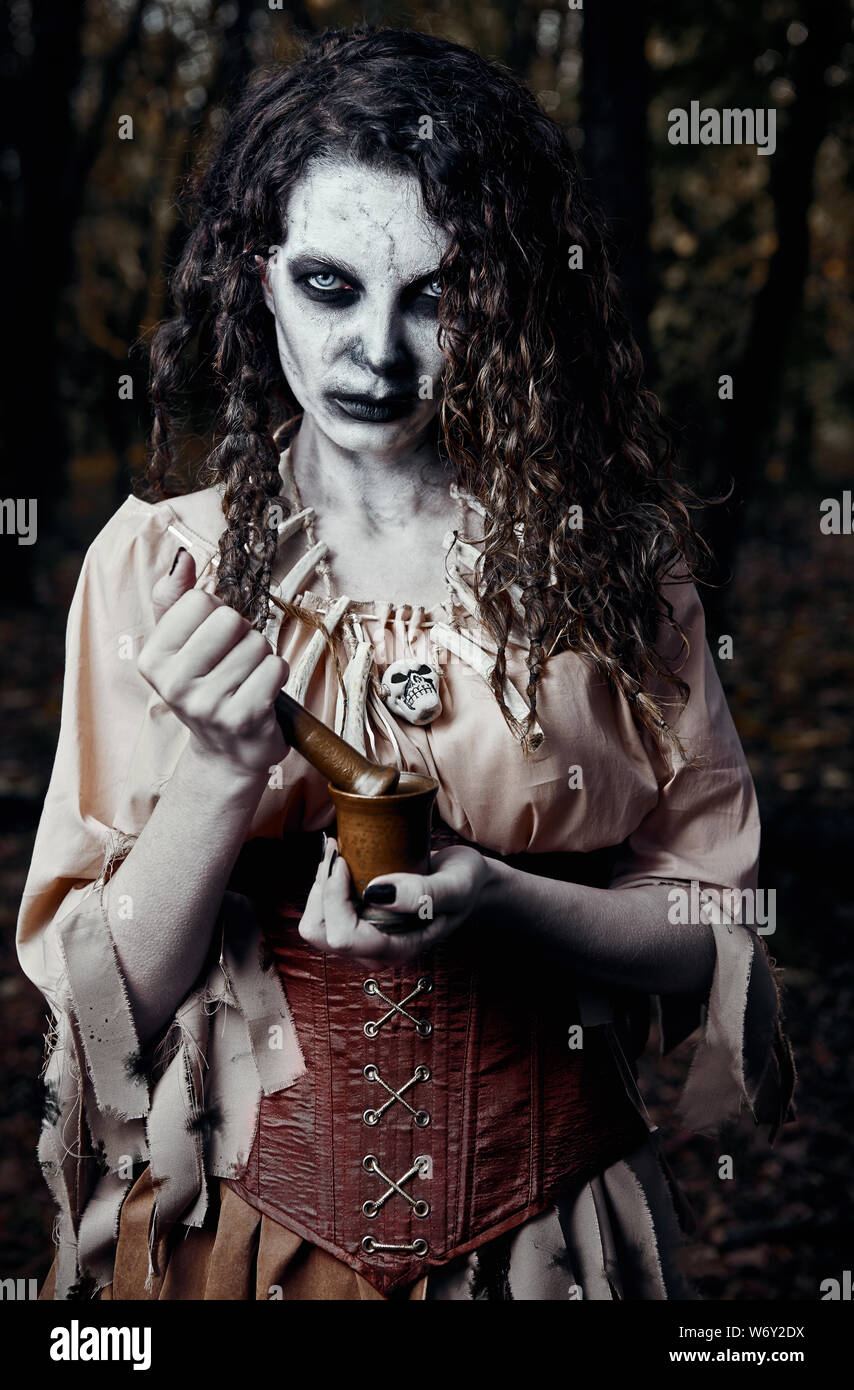 Halloween theme: ugly dread voodoo witch with mortar and pestle. Portrait of the evil hex in dark forest. Zombie woman (undead) Stock Photo