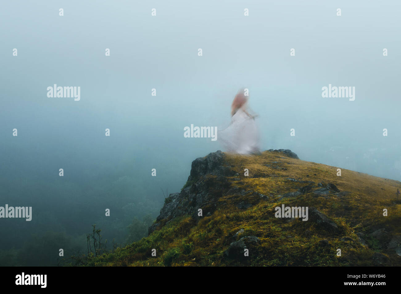 A spooky, blurred, ghostly woman with a white dress, standing on a cliff face looking out on a moody foggy day Stock Photo