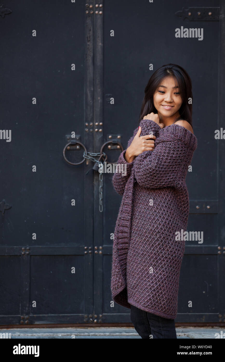 beautiful Asian woman with a purple knitted cardigan hugged herself by the shoulders looking down. Woman standing on the street next to vintage iron d Stock Photo