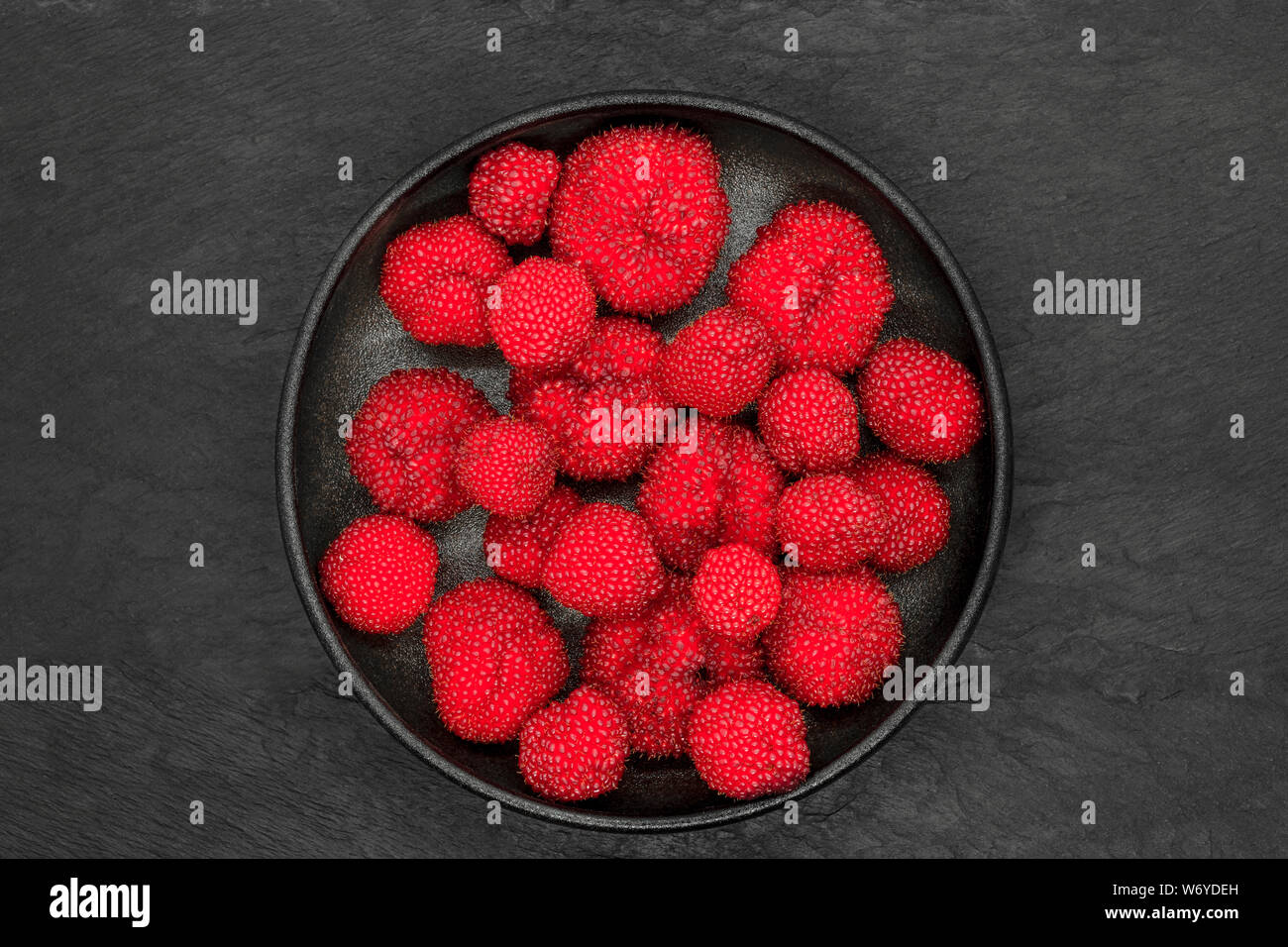Red balloon berries (Rubus Illecebrosus) in round and black iron cast bowl on black stone background Stock Photo