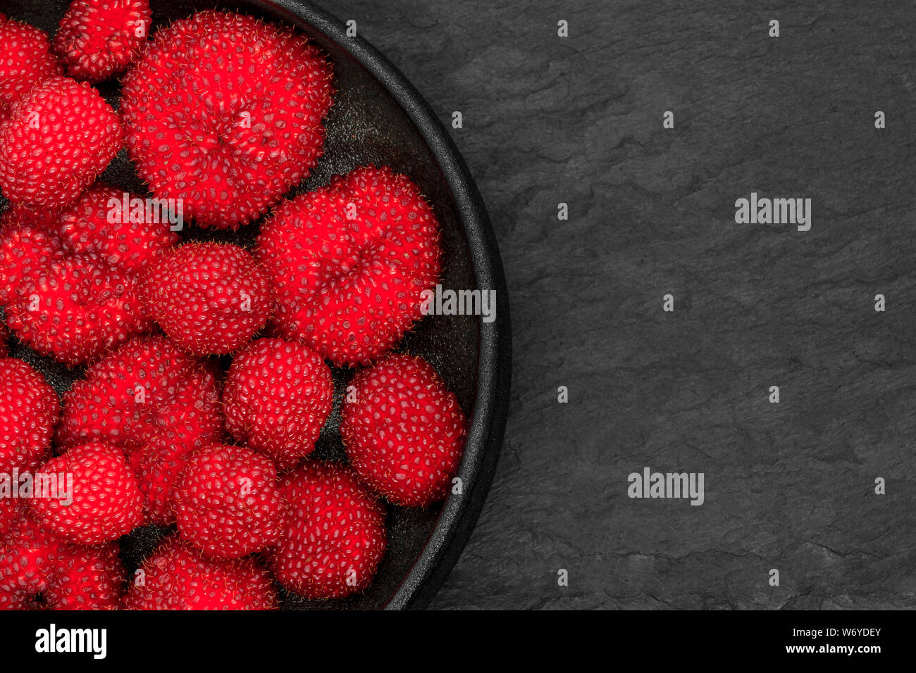 Red balloon berries (Rubus Illecebrosus) in round and black iron cast bowl on black stone background with copy space Stock Photo