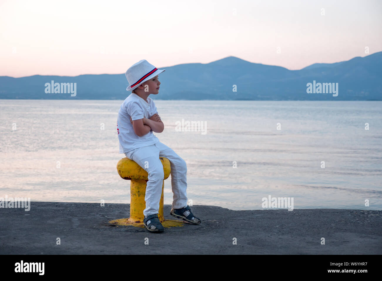 Stylish young hipster sitting on moorage waiting for ship. Little boy dressed in white clothes and hat sitting by the sea. Waiting background. Traveling concept Vacation Holiday Leisuretime Recreation Stock Photo