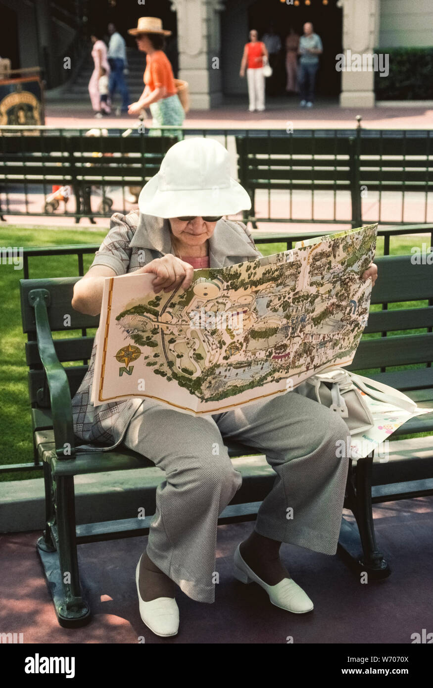 A senior female American visitor to Walt Disney World rests on an outdoor bench while pondering a large illustrated map that shows four theme parks within the world-famous Disney amusement complex near Orlando in Florida, USA. She's trying to decide which one to explore: the Magic Kingdom, Epcot, the Animal Kingdom or Disney's Hollywood Studios. Since opening in 1971, Disney World has become an icon of American culture and is now the most popular vacation resort in the world with more than 52 million visitors annually. Stock Photo