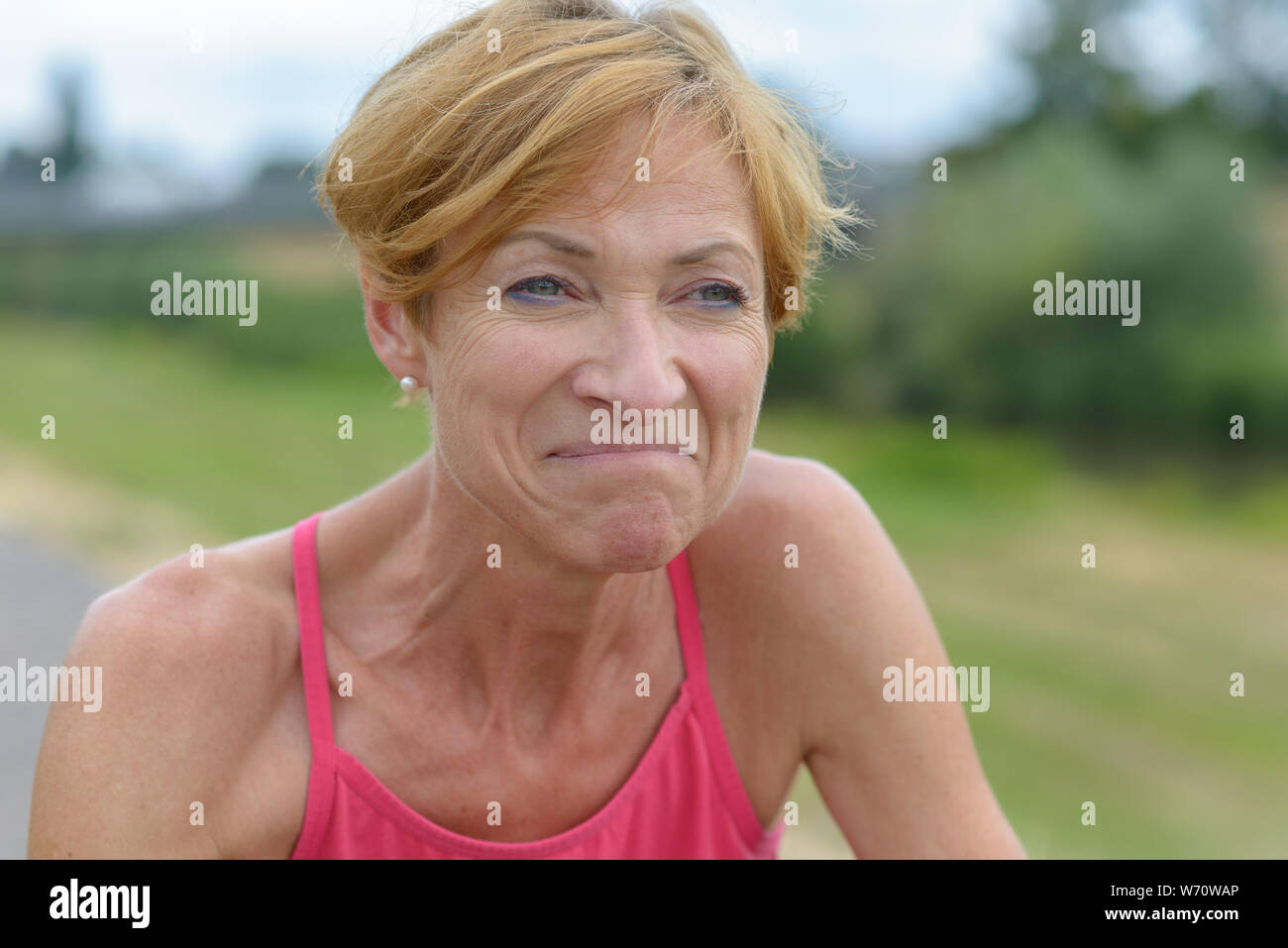 Middle-aged woman grimacing with a wry expression as she stands outdoors in summer in the countryside in a close up portrait Stock Photo