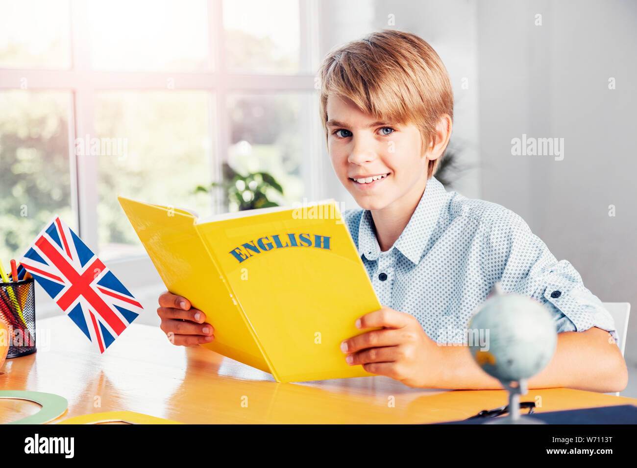 Young blonde smiling boy holding yellow english language book in light classroom Stock Photo