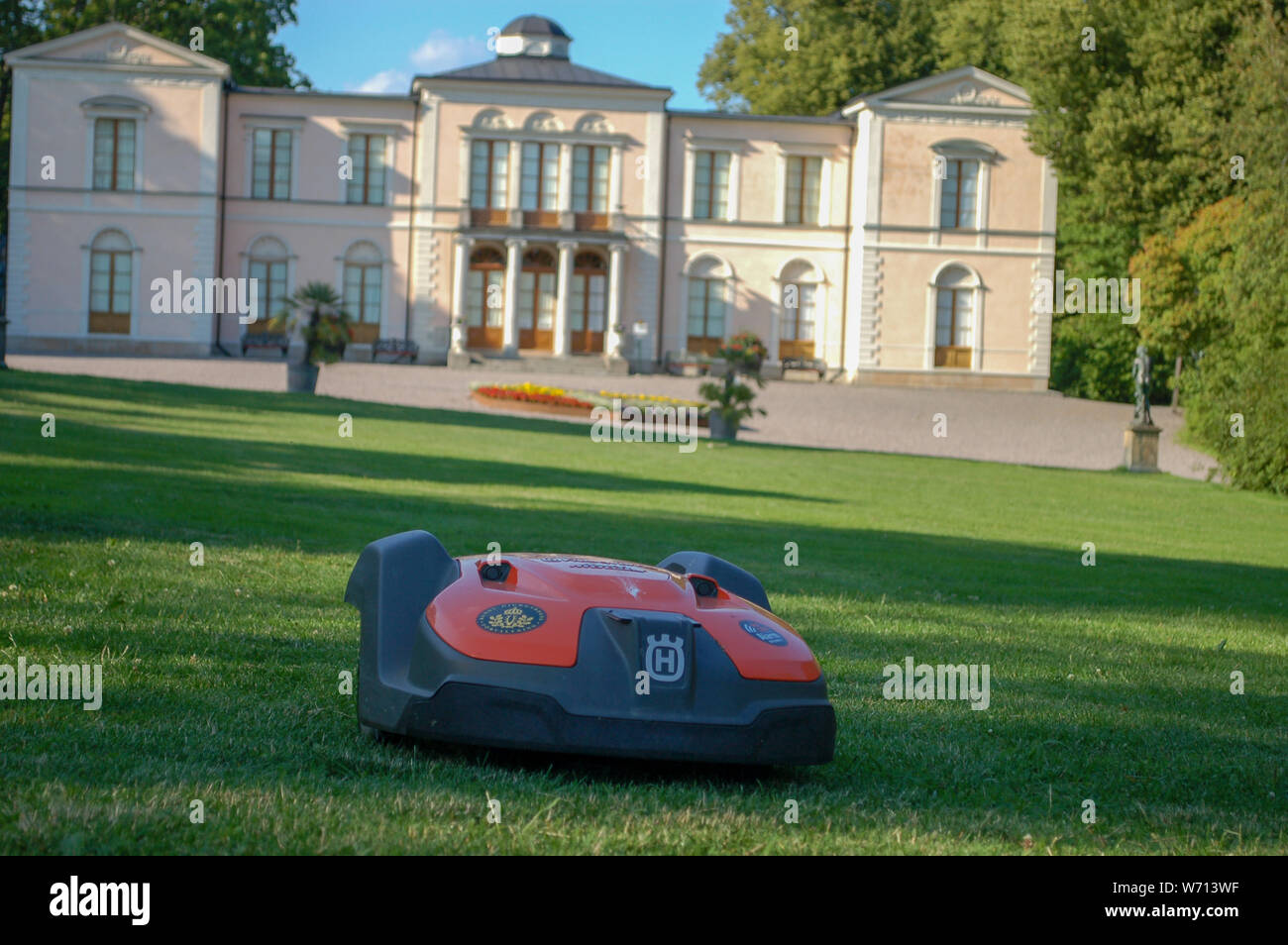 Red Husqvarna robot lawn mower cutting grass on green lawn with castle and blue sky in background, Djurgården, Stockholm, Sweden, Europe Stock Photo