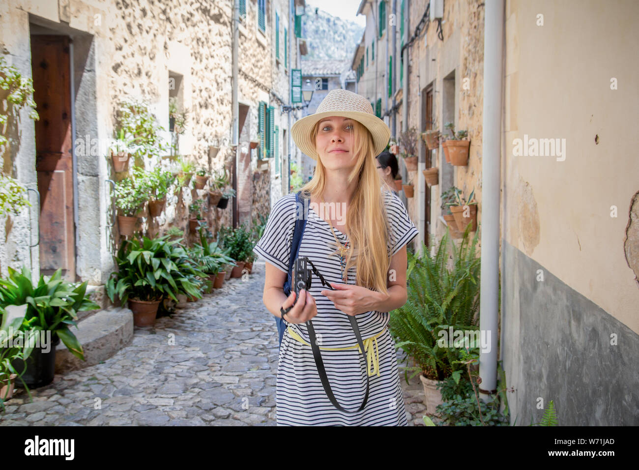 young woman tourist with camera walking down the charming street in mediterranean town in summer Stock Photo