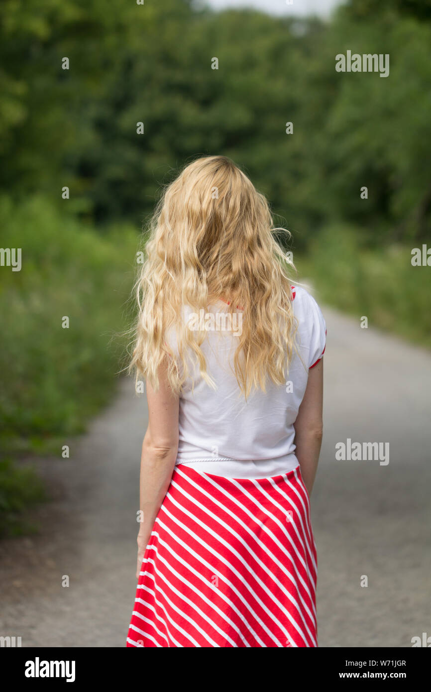 rear view of blonde woman wearing red skirt walking on forest path Stock Photo