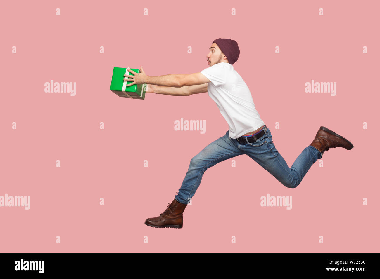 Side view of portrait of crazy bearded young hipster man in white shirt and casual hat jumping, running and hurry up with delivery green present. Indo Stock Photo