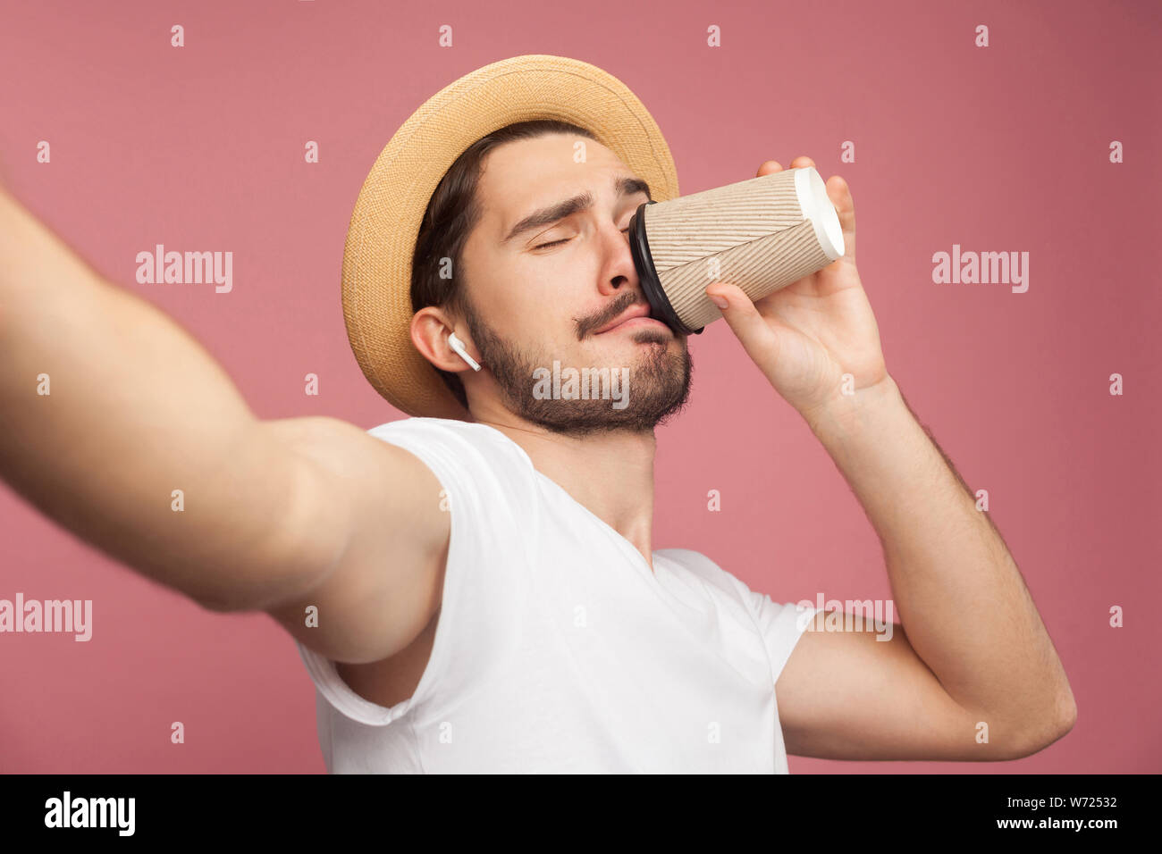 Close up portrait of fashion handsome bearded young hipster blogger in white shirt and casual hat posing and making selfie on phone, tasting coffee. I Stock Photo